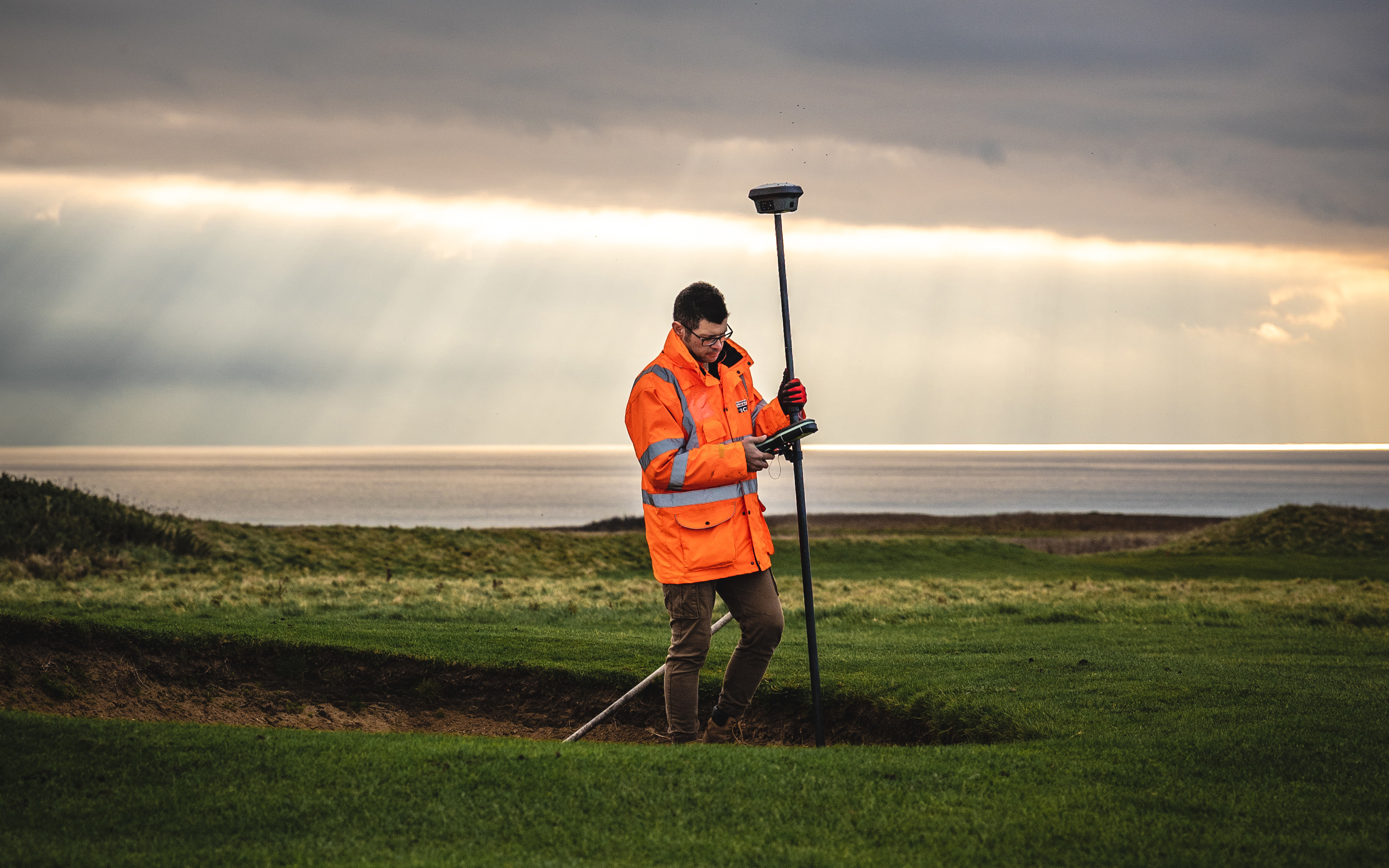 A man stands in a bunker of a golf course holding a GPS receiver. Behind him the sea is visible beneath a dramatic sky.