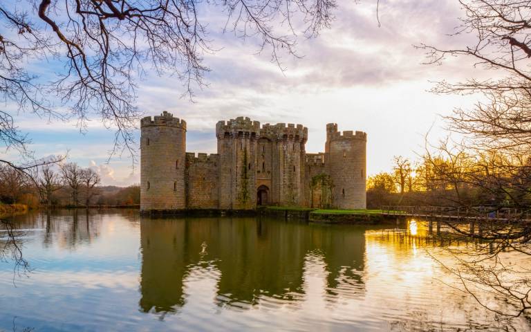 An imposing castle with crenelated walls and towers sits on a large calm moat. Low sun reflects off the water.