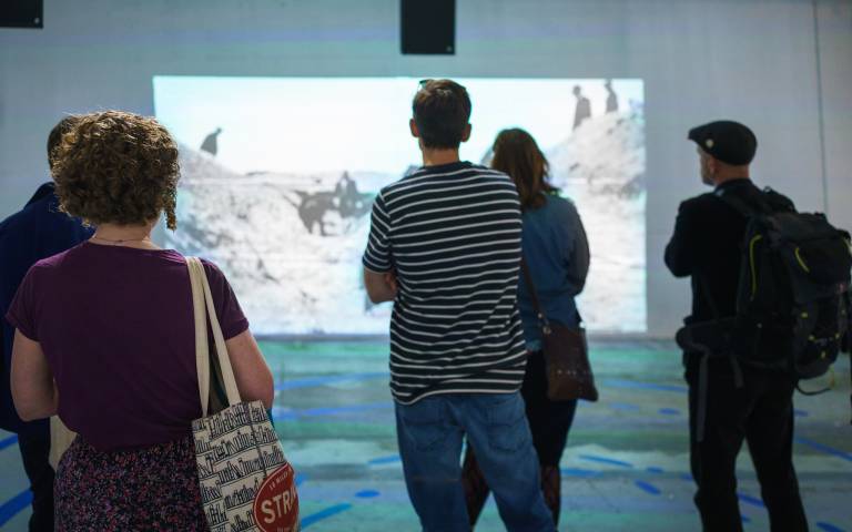 Five people viewed from behind stand looking at projected images of historical archaeological excavations. Blue markings on the floor around them denote the concentric ditch circles of Whitehawk Neolithic Camp.