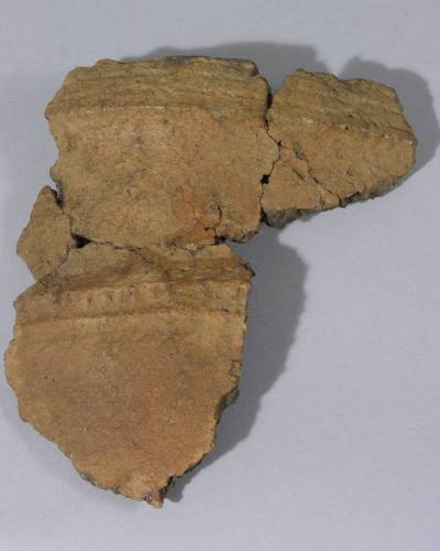 : Four main fragments of a beige ceramic vessel are pieced together. The fragments have some simplistic decoration using grooves and line indentations. The vessel is still incomplete