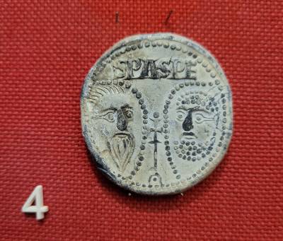 A small piece of circular metal sits on red fabric. The metal is stamped with two faces looking towards each other – both have facial hair, and a prominent nose. Around each face and the circumference of the coin are dots, and between the two faces is a s