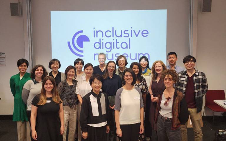 A group of people standing together in a lecture room setting with a project logo on a screen behind them with wording 'Inclusive Digital Museum Innovation'