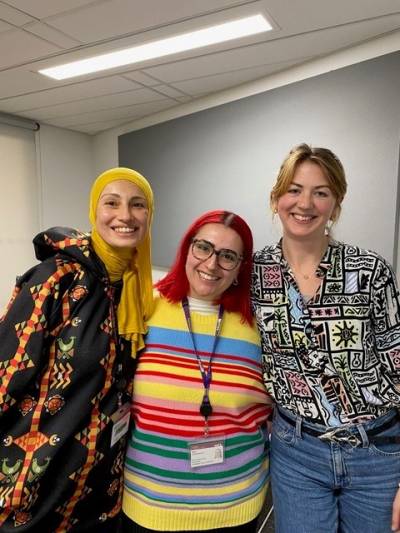 Three women wearing colourful clothes standing together, smiling towards the camera