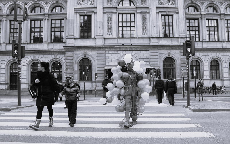 black and white image of a woman and child walking across the road next to a person covered in balloons