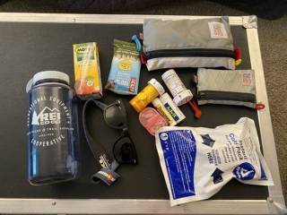 2.	The image shows items that would usually be inside a rucksack. There are two smaller clutches and many separate objects: a water bottle, sunglasses, different containers for medication, instant cold pack, protein bar and packet of juice.