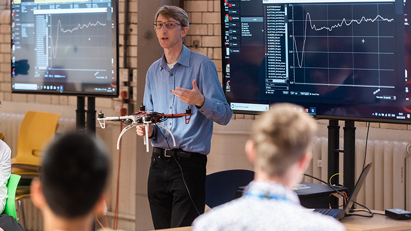 male tutor delivering a lecture with screens displaying graphs and other data representations