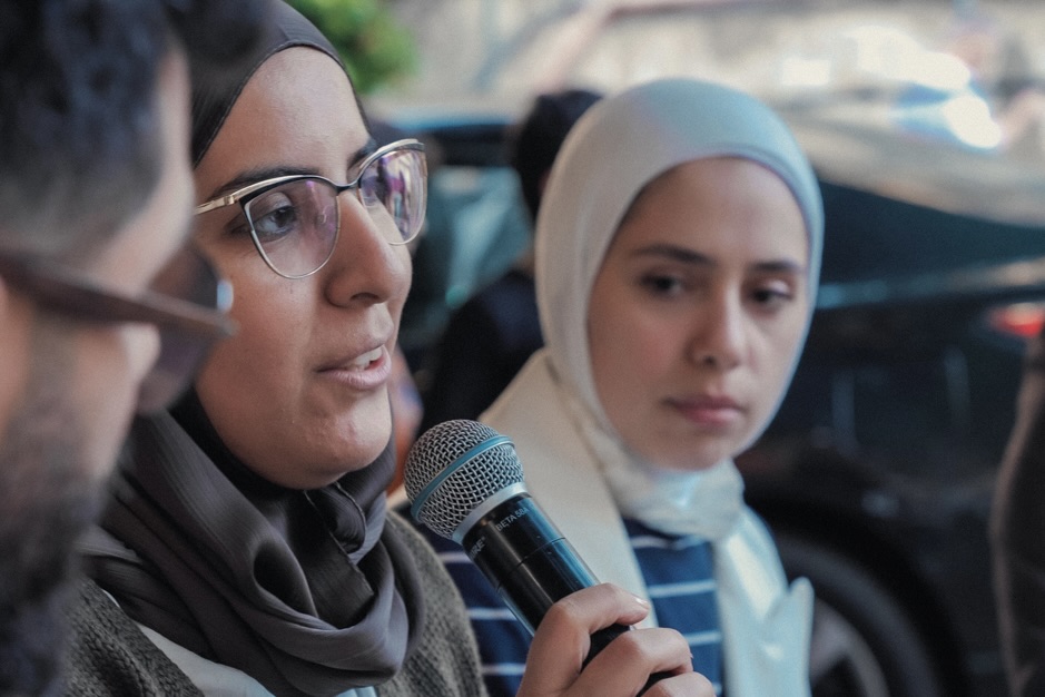 Three people having a discussion. The person in the centre is wearing glasses and a hijab and is speaking into a microphone and looking out of the image towards the person they're answering