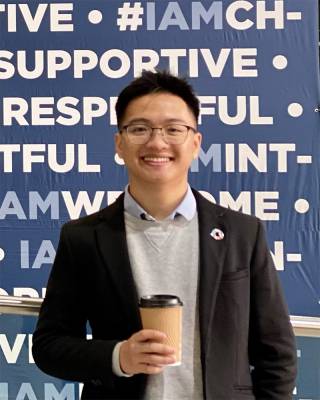 Luong Me stands smiling in front of a banner holding a coffee cup, wearing a blazer, sweater and shirt, and they look happy to be there.