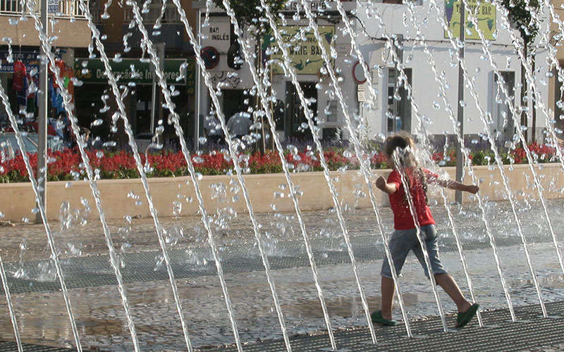 Small child in red t shirt and shorts running through water feature in city