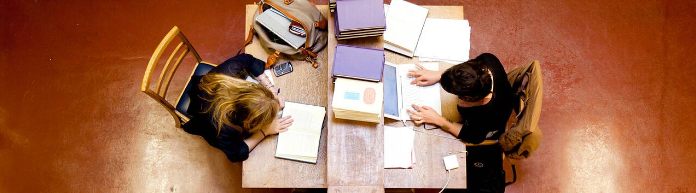 Two students working in the library with books and laptops