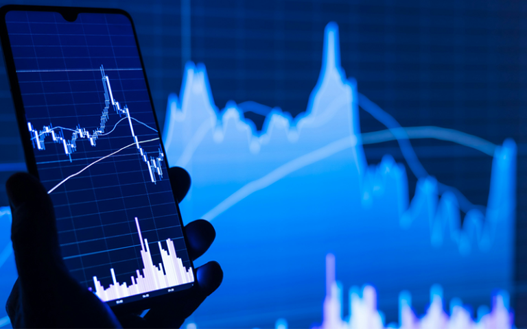 A smartphone screen displays a financial chart with graphs, with a larger similar chart in the background, both showing fluctuating data. The colours are predominantly blue.