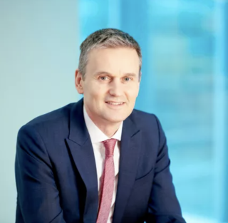 A portrait photograph of Simon. Simon wears a navy blue suit with a pink tie. Simon is fair skinned with grey hair and is smiling. Simon sits in front of a bright blue background