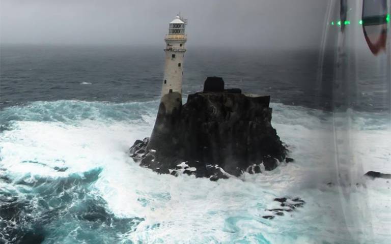 A white lighthouse on black rocks surrounded by waves. 
