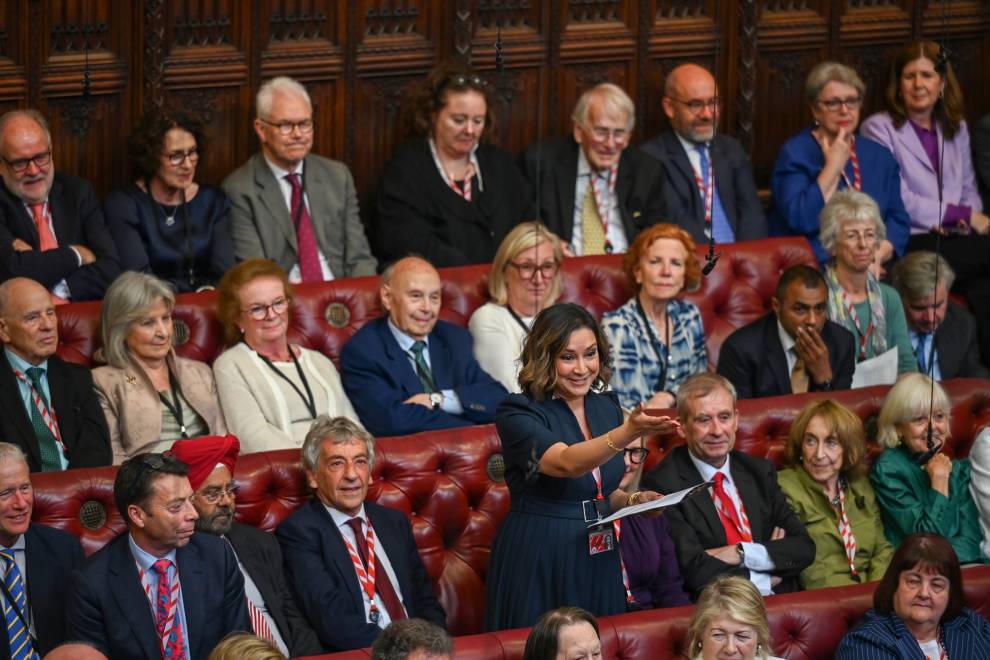 Baroness Hazarika stands in the House of Lords chamber, giving a speech. She is smiling and gesturing towards the other side of the chamber. She is surrounded by other members of the Lords, who are sat on the room’s red benches.