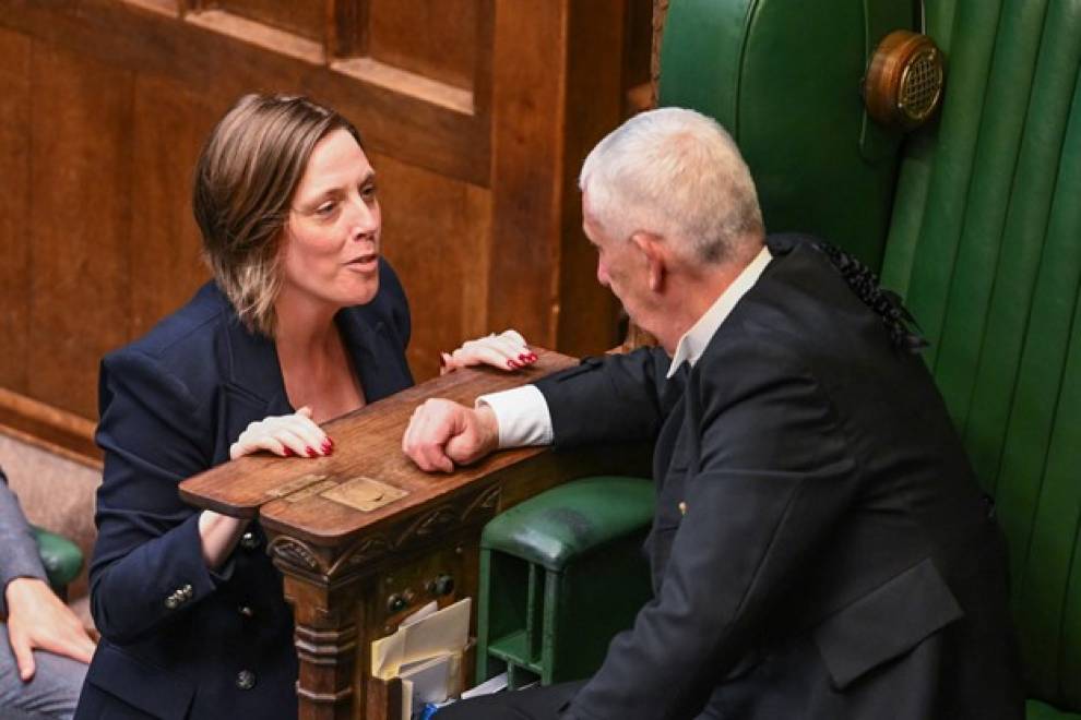 Jess Phillips stands in the House of Commons chamber, hands resting on a wooden frame, beside a large green leather chair. In the chair, back to camera, is Lindsay Hoyle, Speaker of the House of Commons. The two are in conversation. 