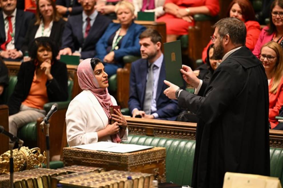 Shabana Mahmood, wearing a hijab and holding a book, stands in the House of Commons chamber, with other MPs watching on. A clerk, dressed in a black gown, is holding up a a green card, which Mahmood is reading from.