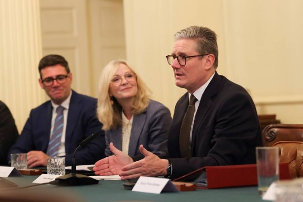 Keir Starmer is shown from the waist up, sat at a table alongside Tracy Brabin and Andy Burnham. Starmer is speaking, with his hands out in front of him. Brabin and Burnham are watching on, smiling.