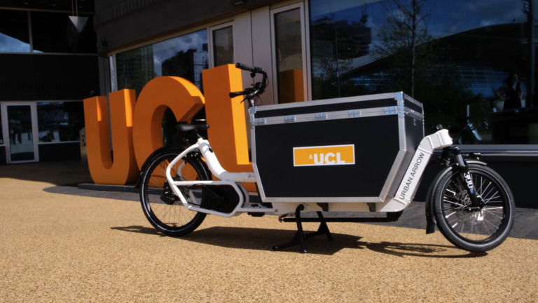 3 wheel black and white cargo bike with the orange UCL logo on it and next to large orange UCL sign