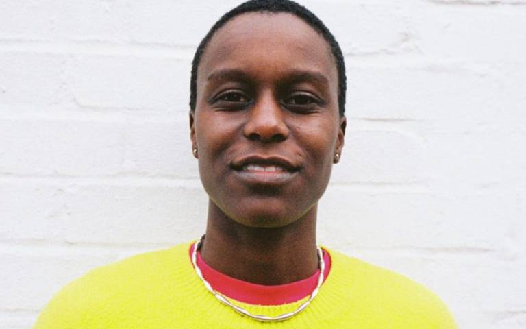 Portrait photo of Remi Graves, a young Black person wearing a bright yellow jumper, smiling slightly and standing against a white wall