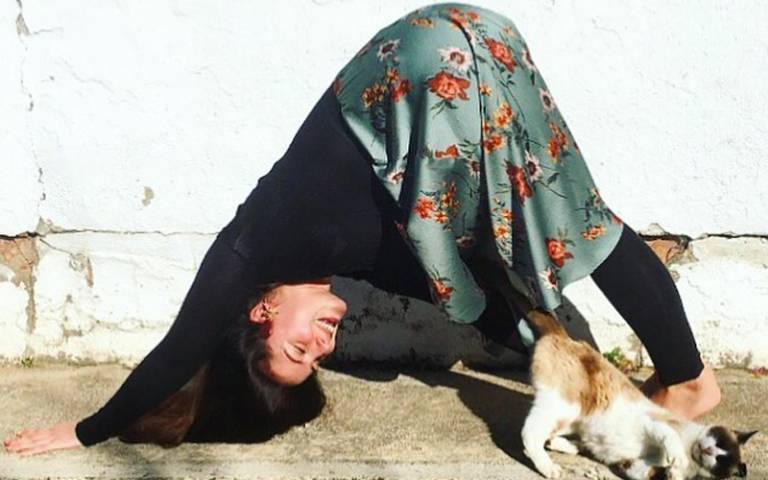 A woman wearing a black top, floral skirt and black leggings holds a downward dog yoga pose outside in the sun, with a cat stretching next to her in a similar pose
