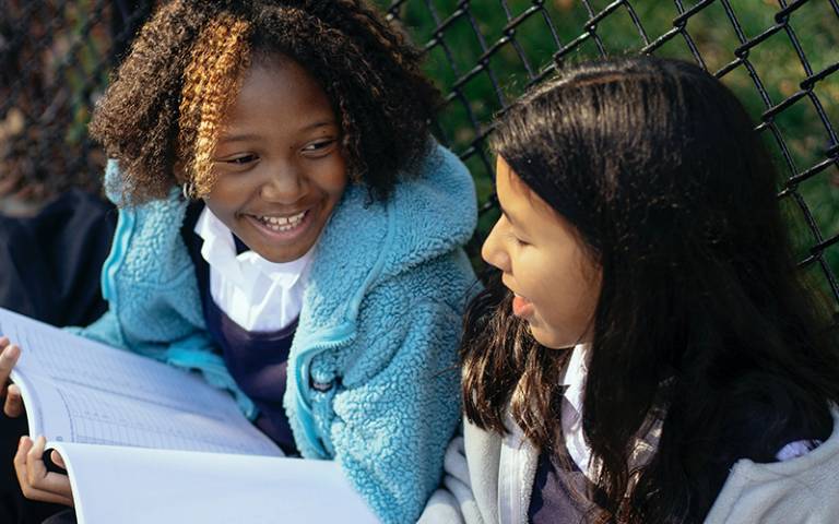 Two school girls are sitting outside by a chain link fence on a sunny day with exercise books, smiling at each other.