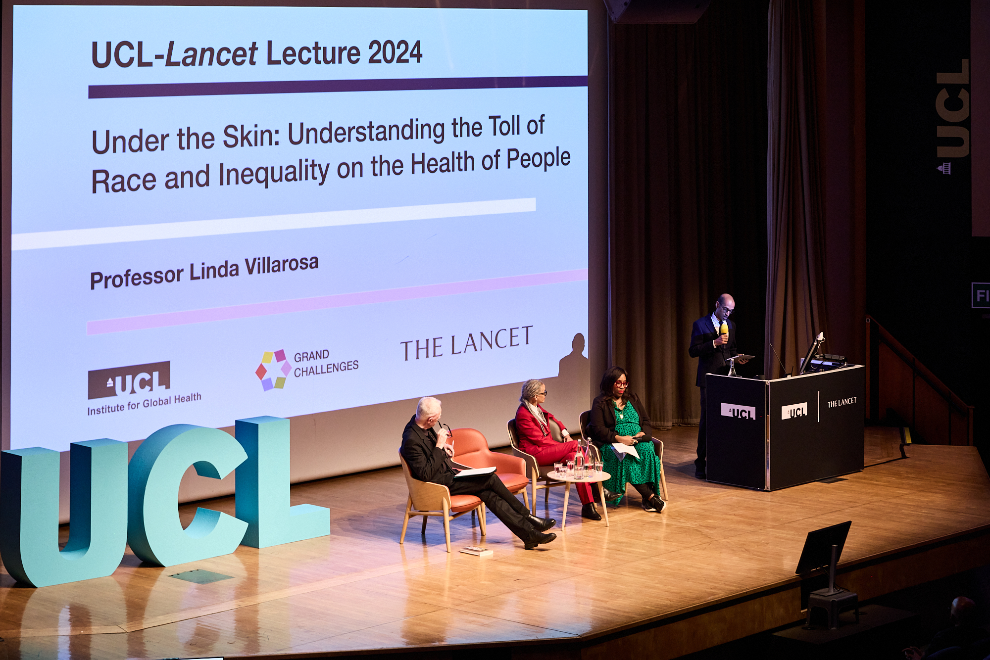 4 speakers sitting in chairs on stage with large UCL letters next to them