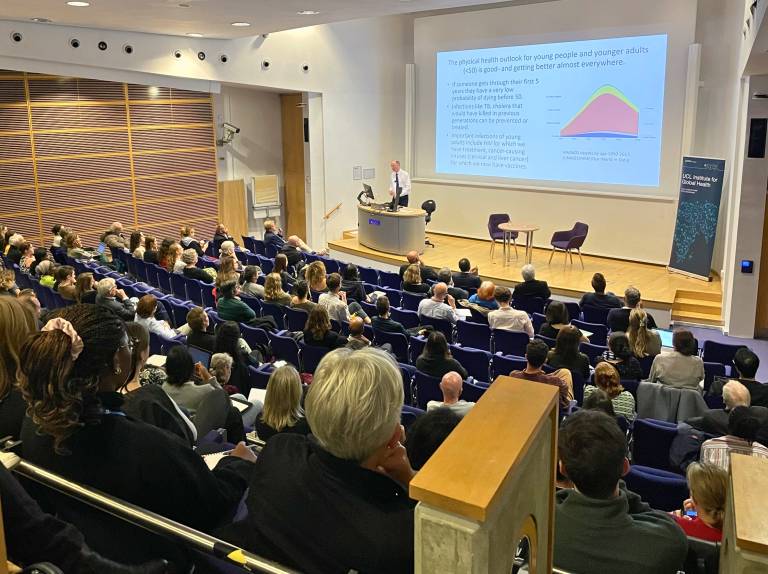 Professor Chris Whitty delivering a seminar on stage to a lecture theatre full of people.