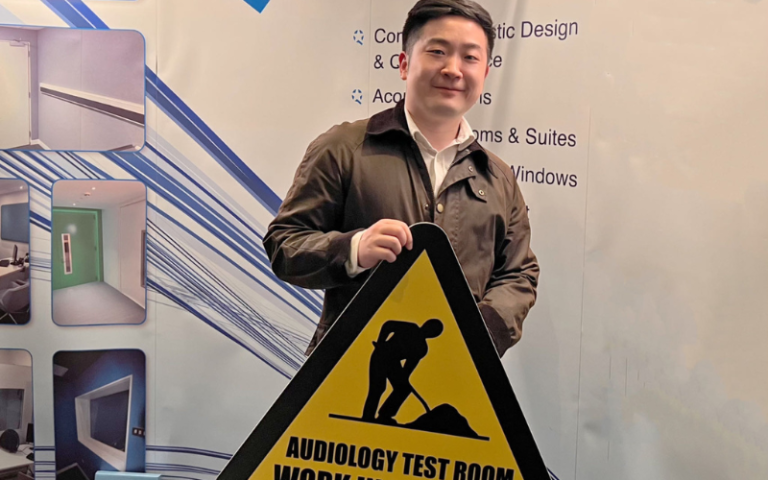 Dr Jinke Chang from UCL Medical Sciences holding a 'Work in Progress' sign at the British Society of Audiology conference. The background features promotional material about acoustic design and audiology test rooms, highlighting the collaborative researc