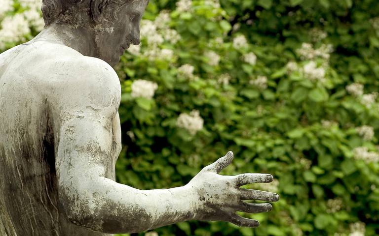 Image of a statue of a man against a background of leaves. The statue is seen in profile and has its hand outstretched.