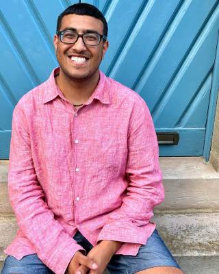 A man in a pink shirt smiles in front of a blue background