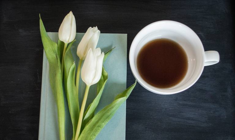 Looking from above down on a wooden table. On the right a mug with some tea in it. On the left, a book with a pale blue cover on top of which some white tulips are placed