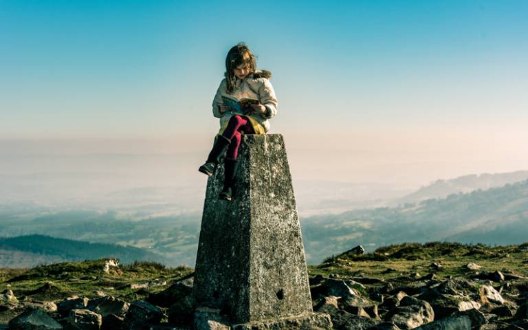 Child sitting on a tall block at the top of a mountain and reading. Image: Carl Jorgensen via Unsplash