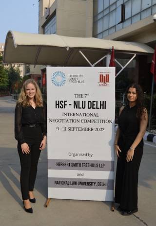 Nikki Marashi and Mette Gutjahr standing beside a banner which text: "HSF-NLU Delhi International Negotiation Competition 9-11 September 2022"