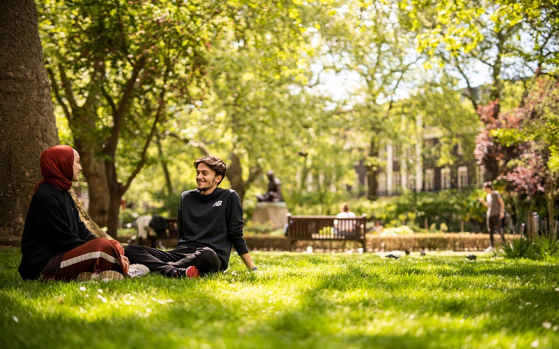 Two people are sitting on grass in Tavistock Square and are talking. In the background are trees with leaves, a bench, a statues and some buildings.