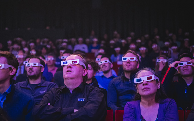 A group of people wearing 3D glasses and watching something with their faces to the camera