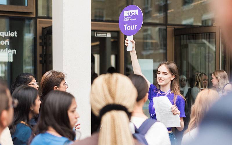 UCL student holds up a campus tour placard surrounded by prospective student guests