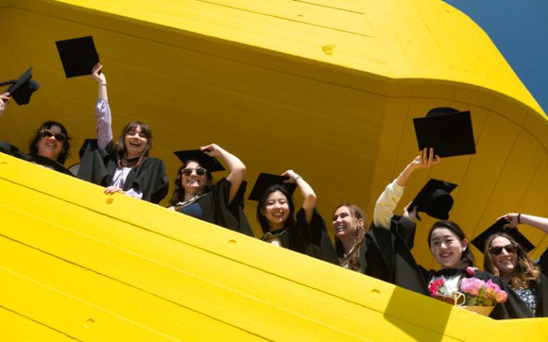 Seven graduates in academic dress standing on a bright yellow staircase at the Royal Fesitival Hall, looking happy and waving their caps in the air.