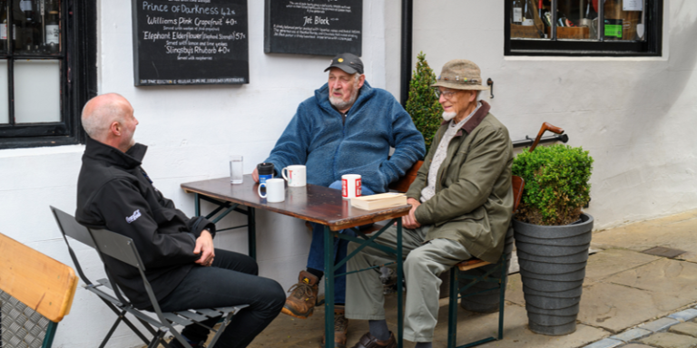 Three older men sitting at a table with mugs of tea outside a pub in England