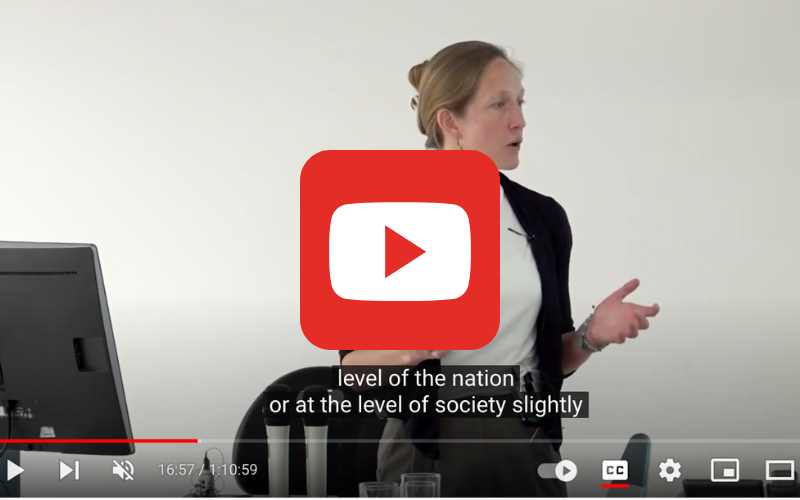 The YouTube play button is placed ontop of a screen grab from a lecture given by Lucy Barnes. Lucy is speaking to an audience