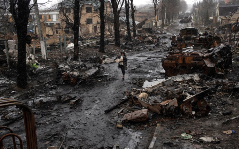 A person in a rain coat with the hood up walks through a street covered in building rubble and burnt out vehicles. Some of them are millitary vehicles. They are carrying a small cardboard box.