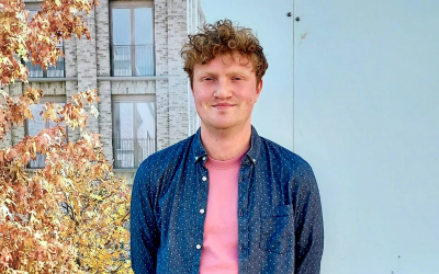 Josh stands outside by an tree with orange leaves. He is wearing a pink tshirt with a dark blue shirt with small white dots on top of it. 