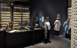 Students look at the records exhibition at International Committee of the Red Cross Museum. There are cabinets from floor to ceiling filled with old paper records in a darkly light room