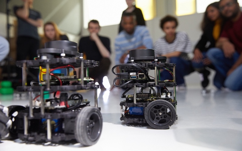 Two small wheeled remote control vehicles on floor with group of students in background