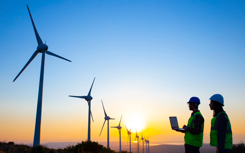 Background sunset over a windfarm. Two workers look on, one is holding a laptop