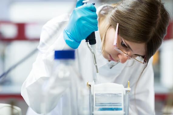 Female student wearing a lab coat and gloves, carefully using a tool in a biology lab. 