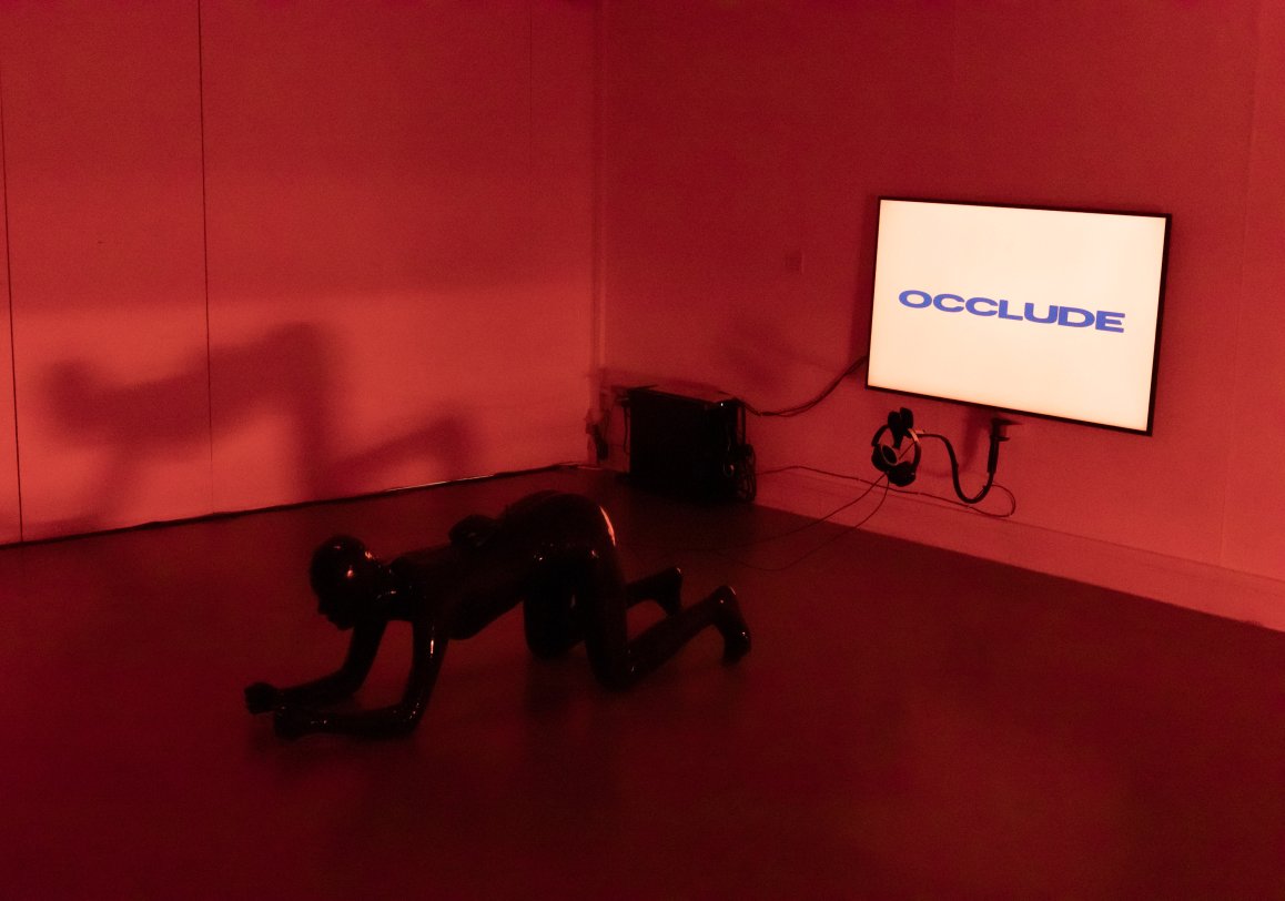 Installation photo of red room, prone black figure/stool and screen on wall with the words "Occlude"