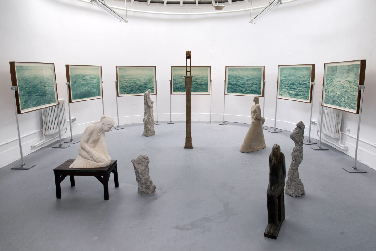 Installation photo of figurative sculpture in the foreground, seascape drawings in the background