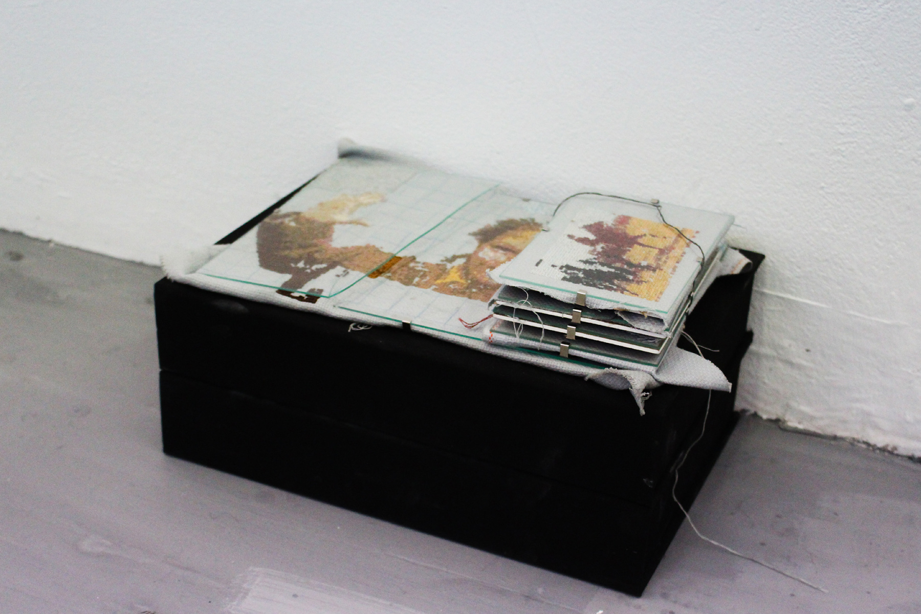 Installation photo of archival boxes, archival material, clip frames, broken glass, embroidery thread, cotton fabric.