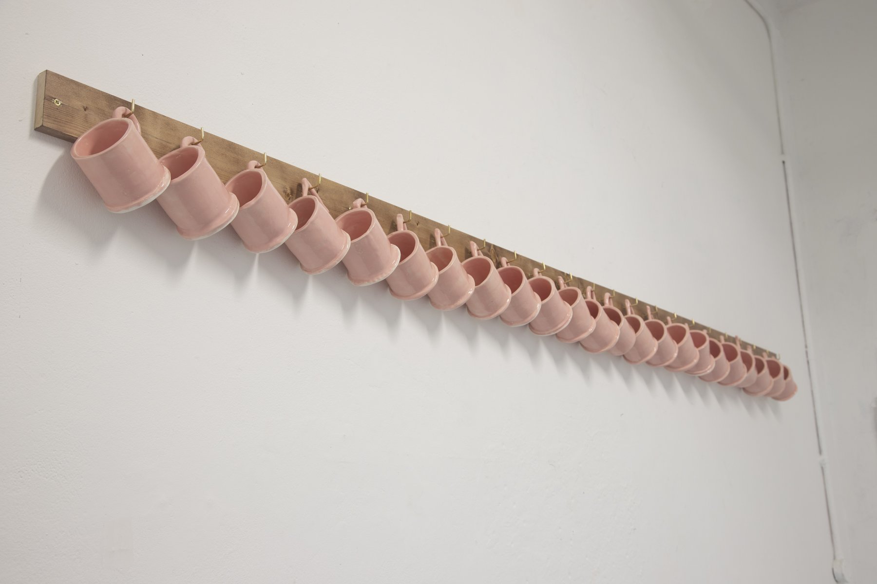 Row of pink earthenware mugs hanging on hooks on wooden rail.