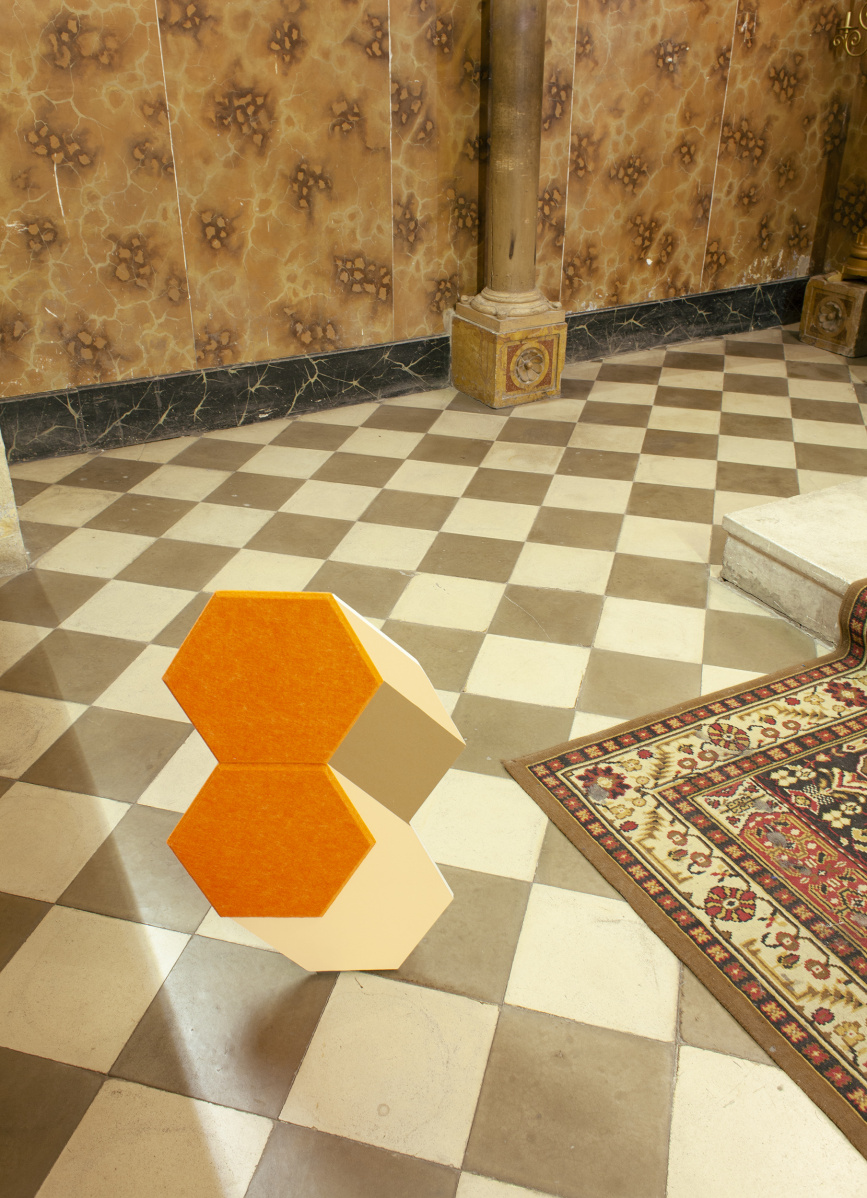 Two 3-d hexagonal shapes, one on top of the other on a tiled floor in a room with pillars against the wall. Part of a decorative rug can be seen in the front corner of the picture.